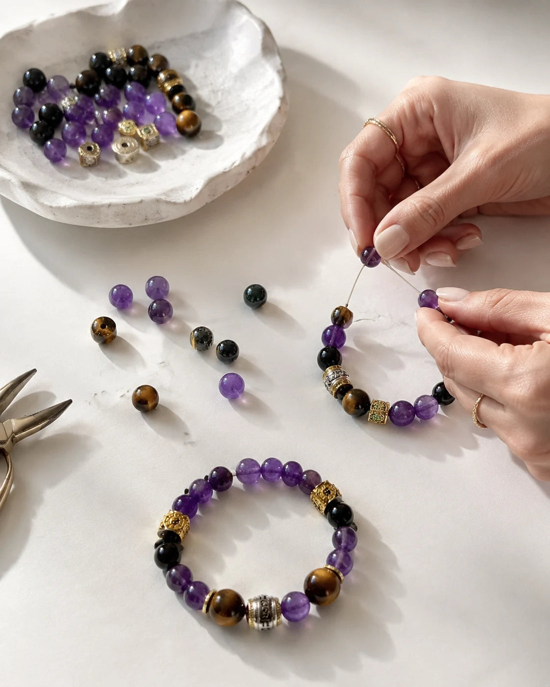 Hands pausing during bracelet assembly with loose beads, focal pieces, and tools arranged on a bright work surface